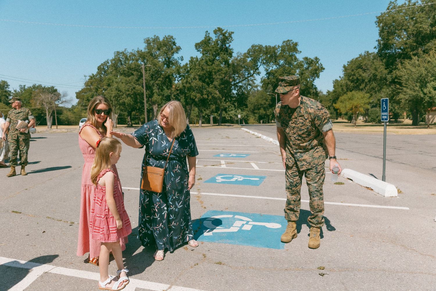 Marine in uniform gestures to family with young girl in parking lot during military homecoming ceremony.