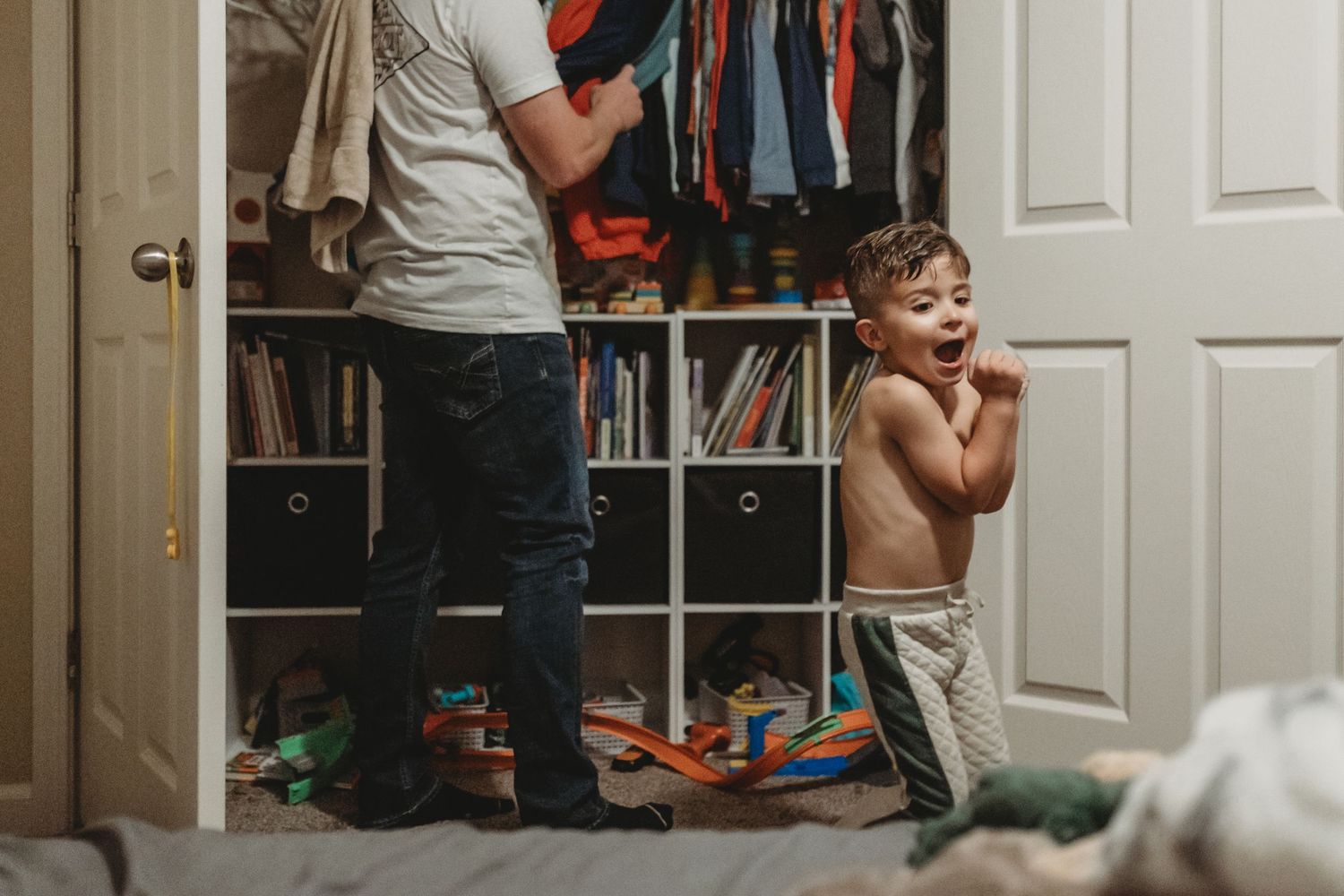 A playful scene in a closet room with a small child and someone looking through hanging clothes.