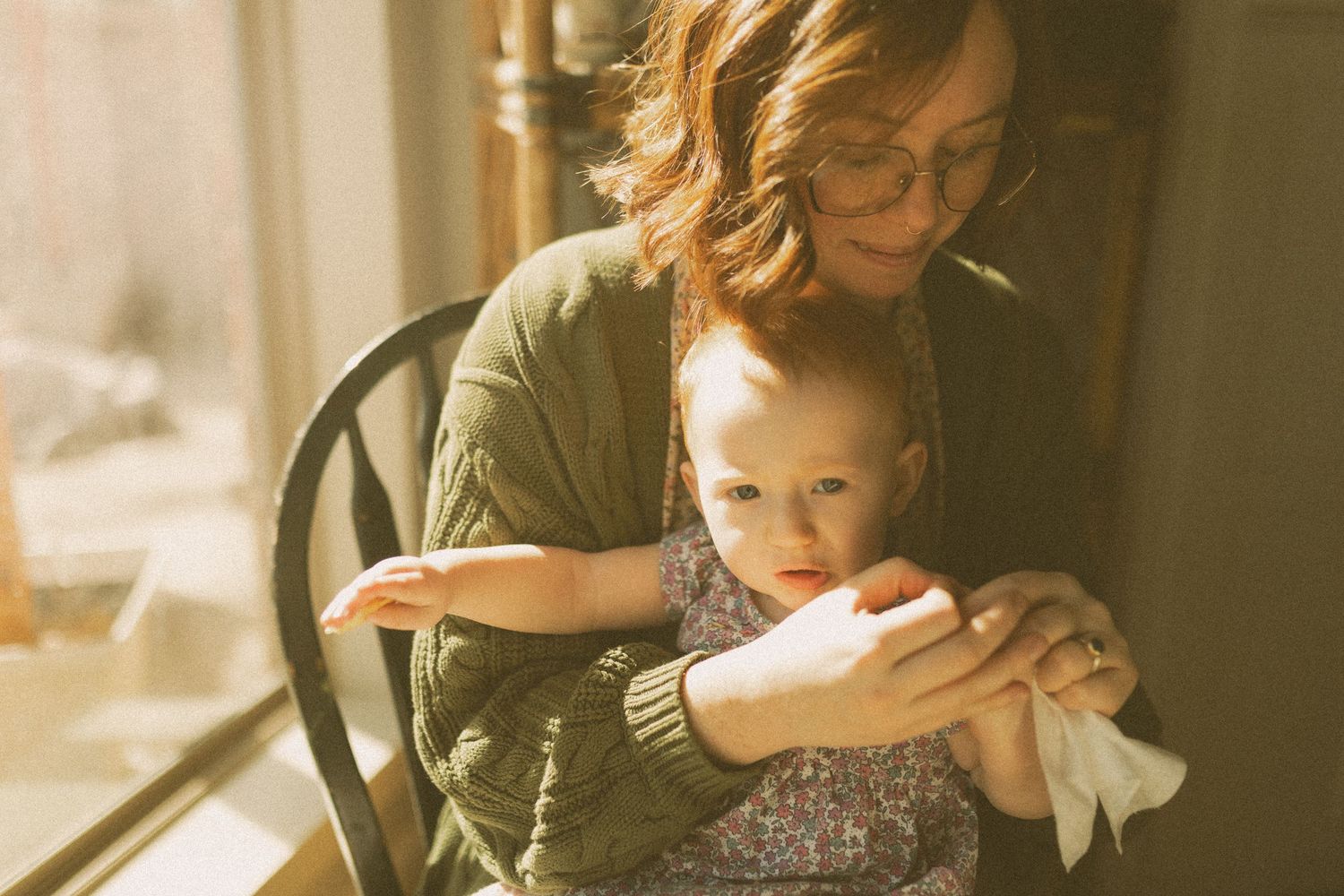A parent lovingly cradles and cleans her baby's nails in soft, natural window light at home.