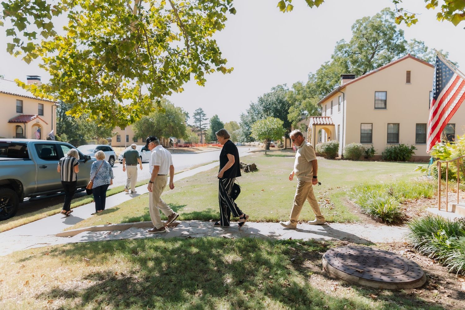 Group of people walking on a sunny sidewalk near a yellow house with an American flag outdoors.
