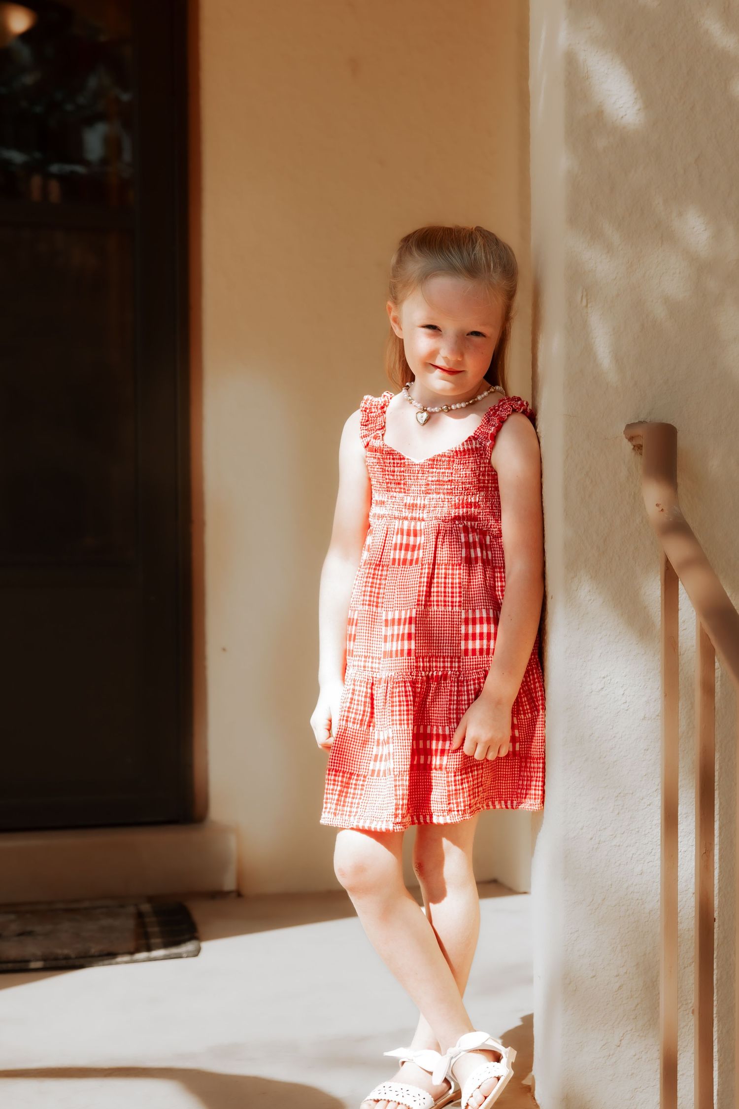 Young girl in red gingham dress smiling against a sunlit wall wearing white sandals and a necklace.