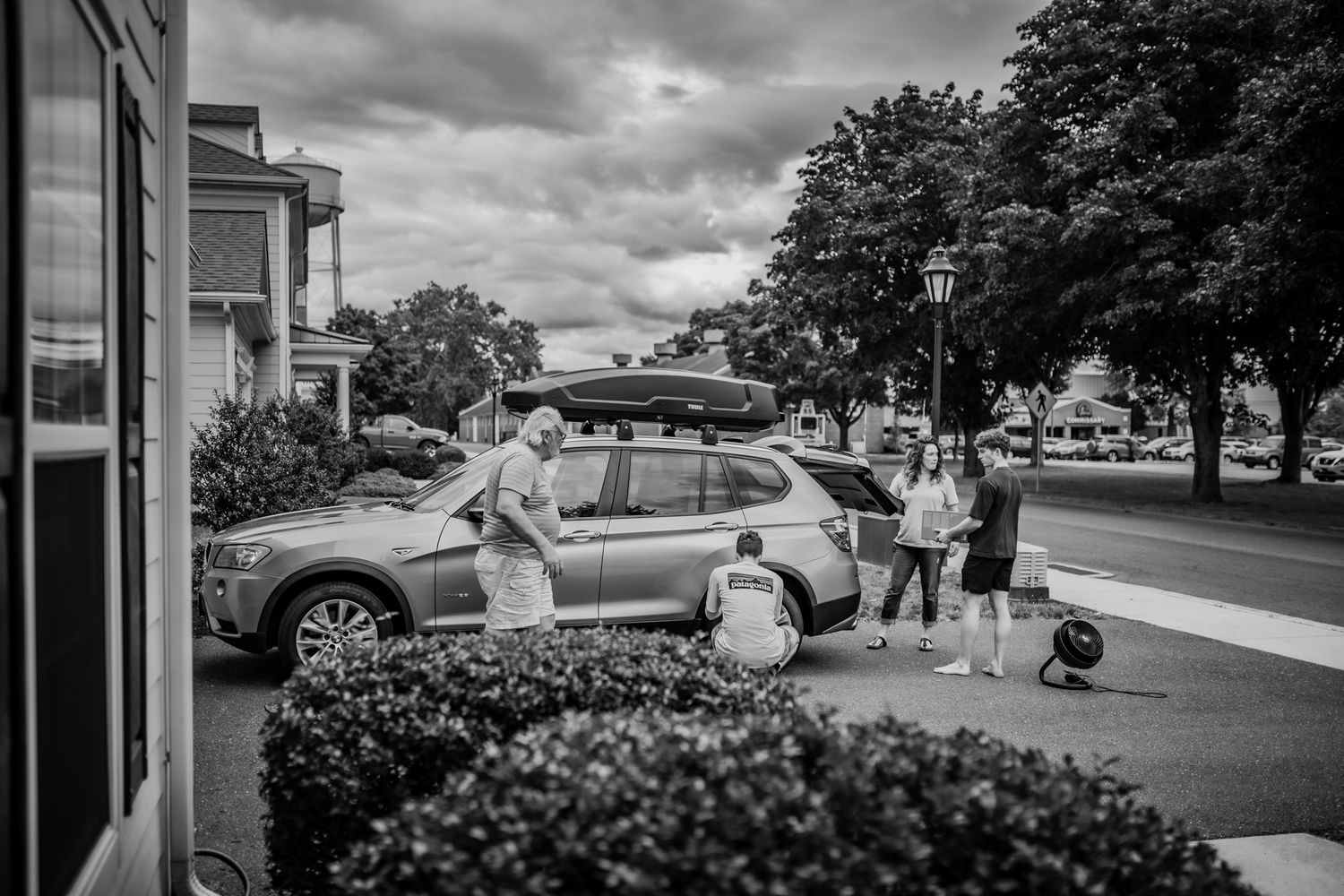 Black and white photo of group loading luggage onto parked car in residential area. In-Home Photography Liz Perez