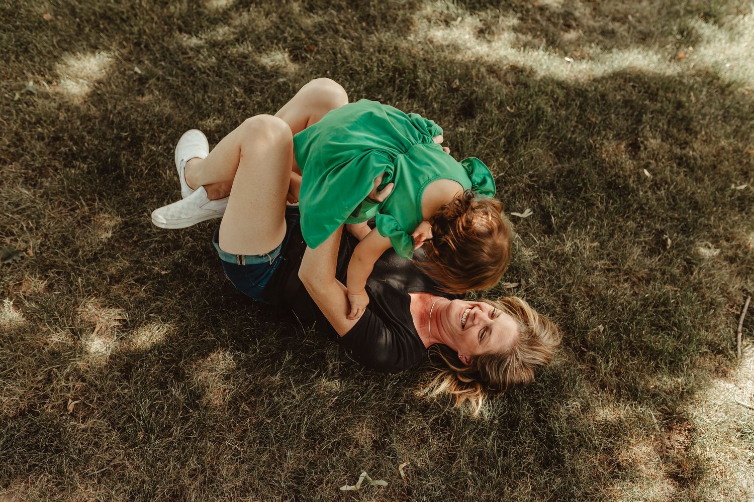 A mother and child laughing and playing together on green grass in a sunny park.