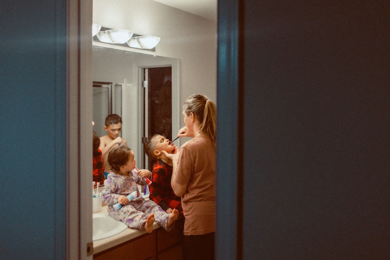 Mother brushing young child's teeth in bathroom mirror reflection at night.