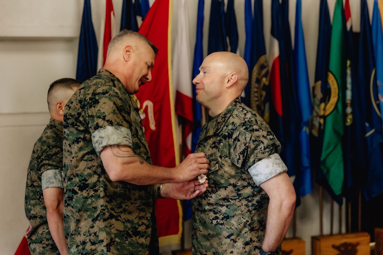 Two U.S. Marines in camouflage uniforms shake hands during a military ceremony with colorful flags in the background.
