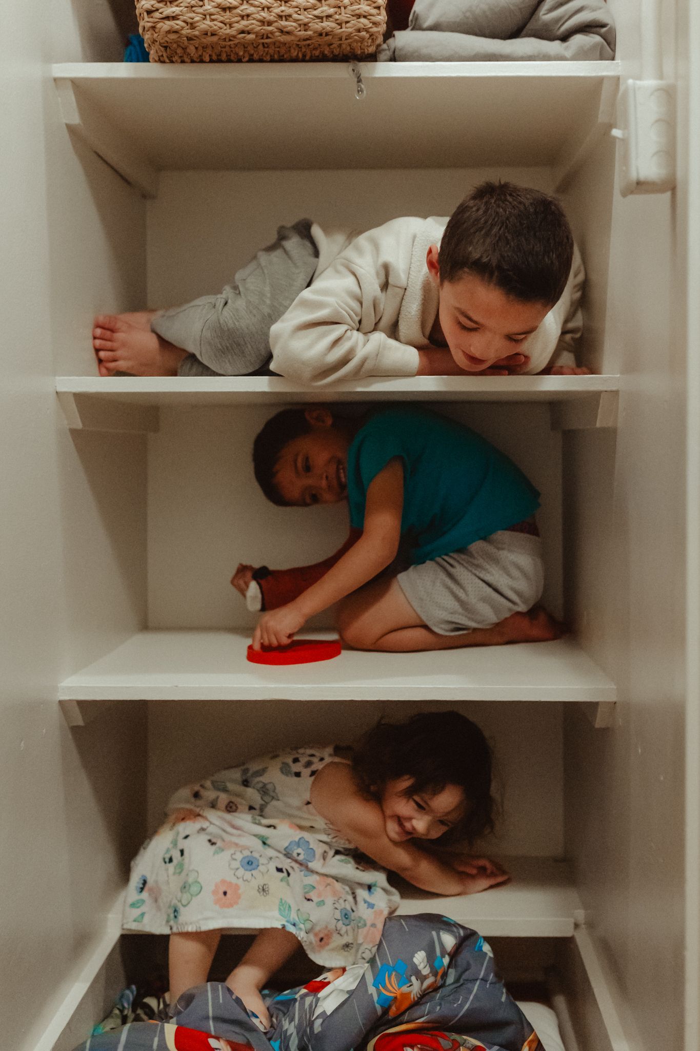 Children lie playfully on white shelves in a closet during what appears to be a fun hide and seek game.