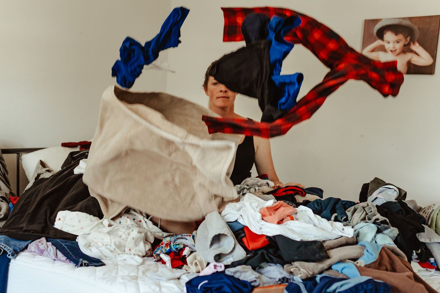 Person tossing clothes onto a large messy pile of laundry on a bed in a cluttered bedroom.