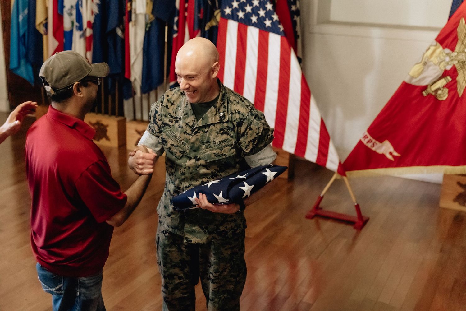 Marine in camouflage uniform shakes hands with civilian while holding folded American flag indoors.