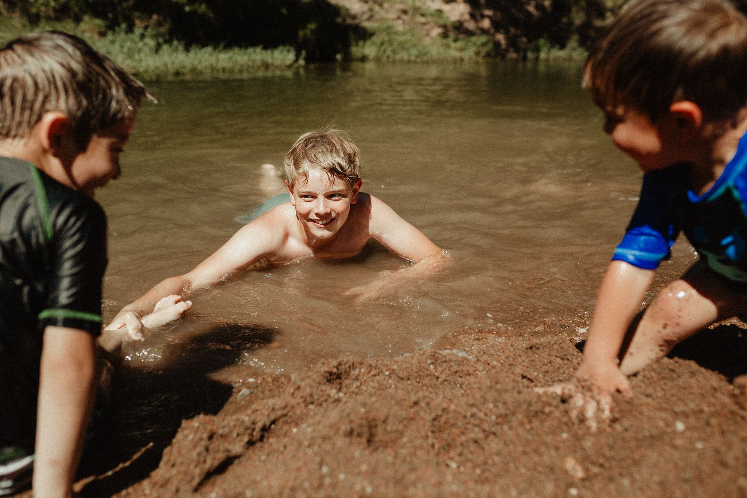 Kids playing in muddy water by a riverbank on a sunny day.