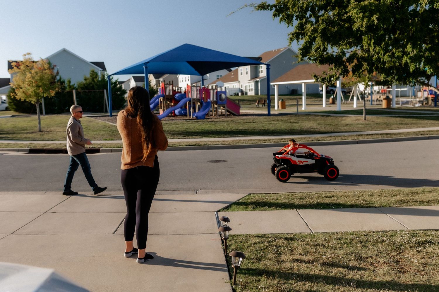 People watching a child ride a toy vehicle on a suburban sidewalk.