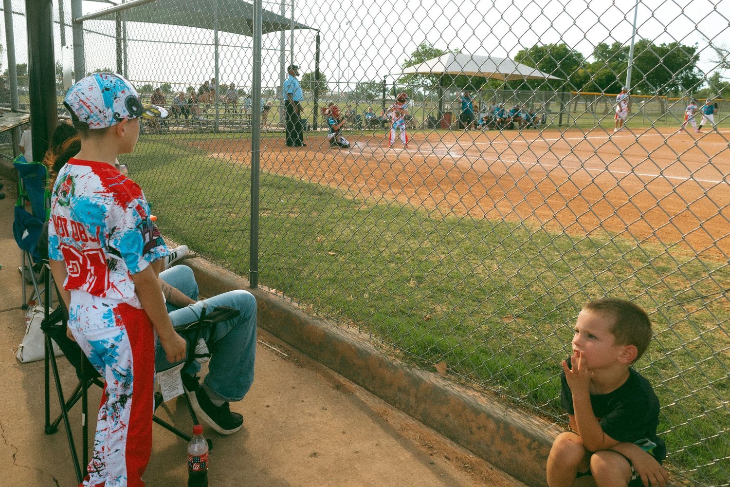 Two children watch a youth baseball game from behind a chain-link fence at a sunny outdoor ballpark.