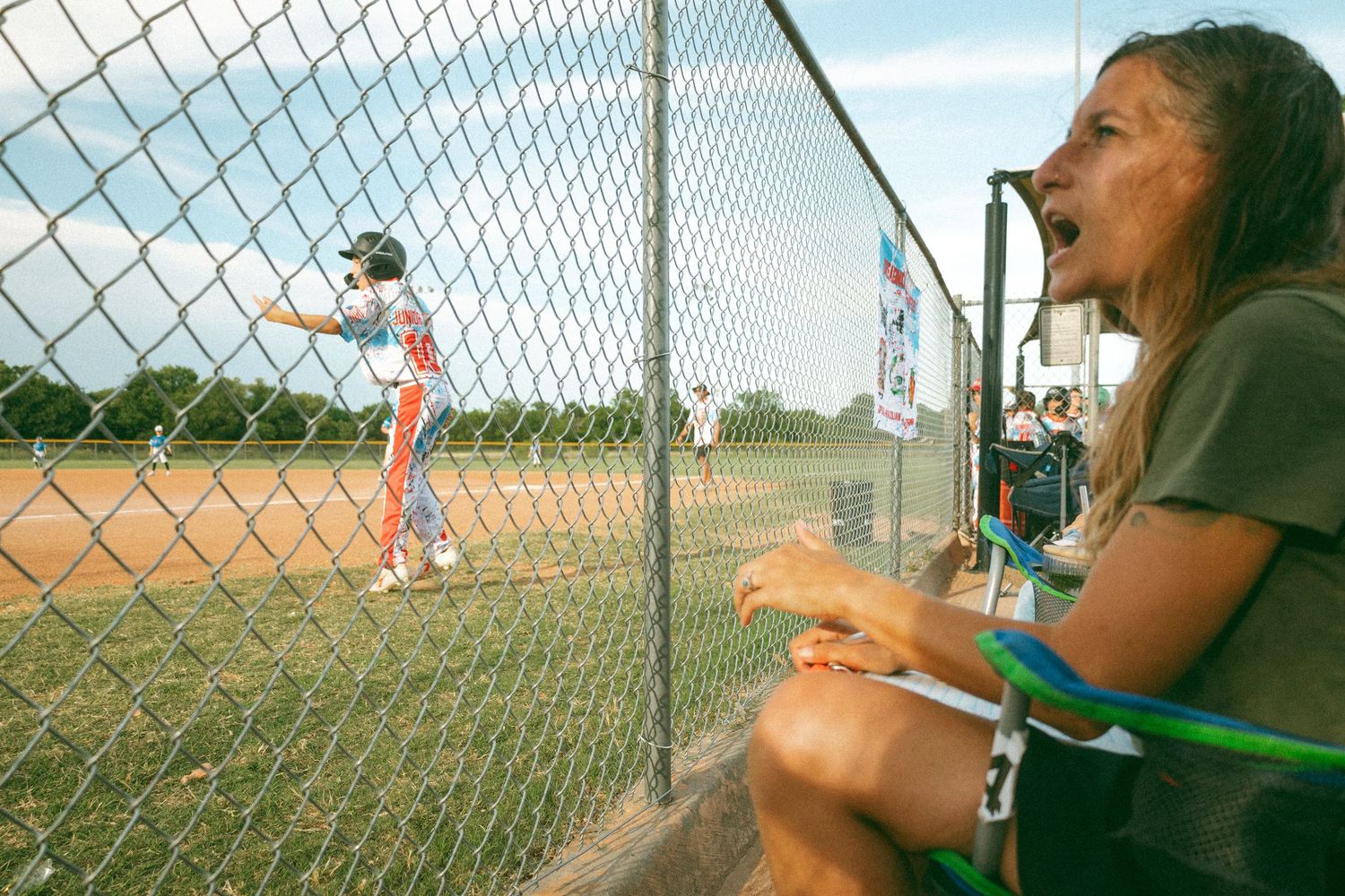 Woman cheering enthusiastically at a youth softball game through a chain-link fence on a sunny day.