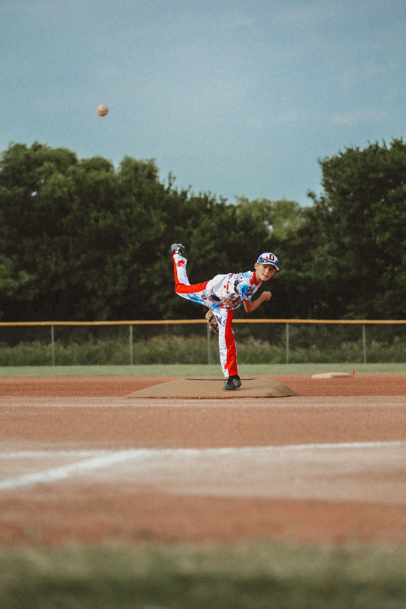 Young baseball pitcher in red, white, and blue uniform mid-throw on mound during little league game.