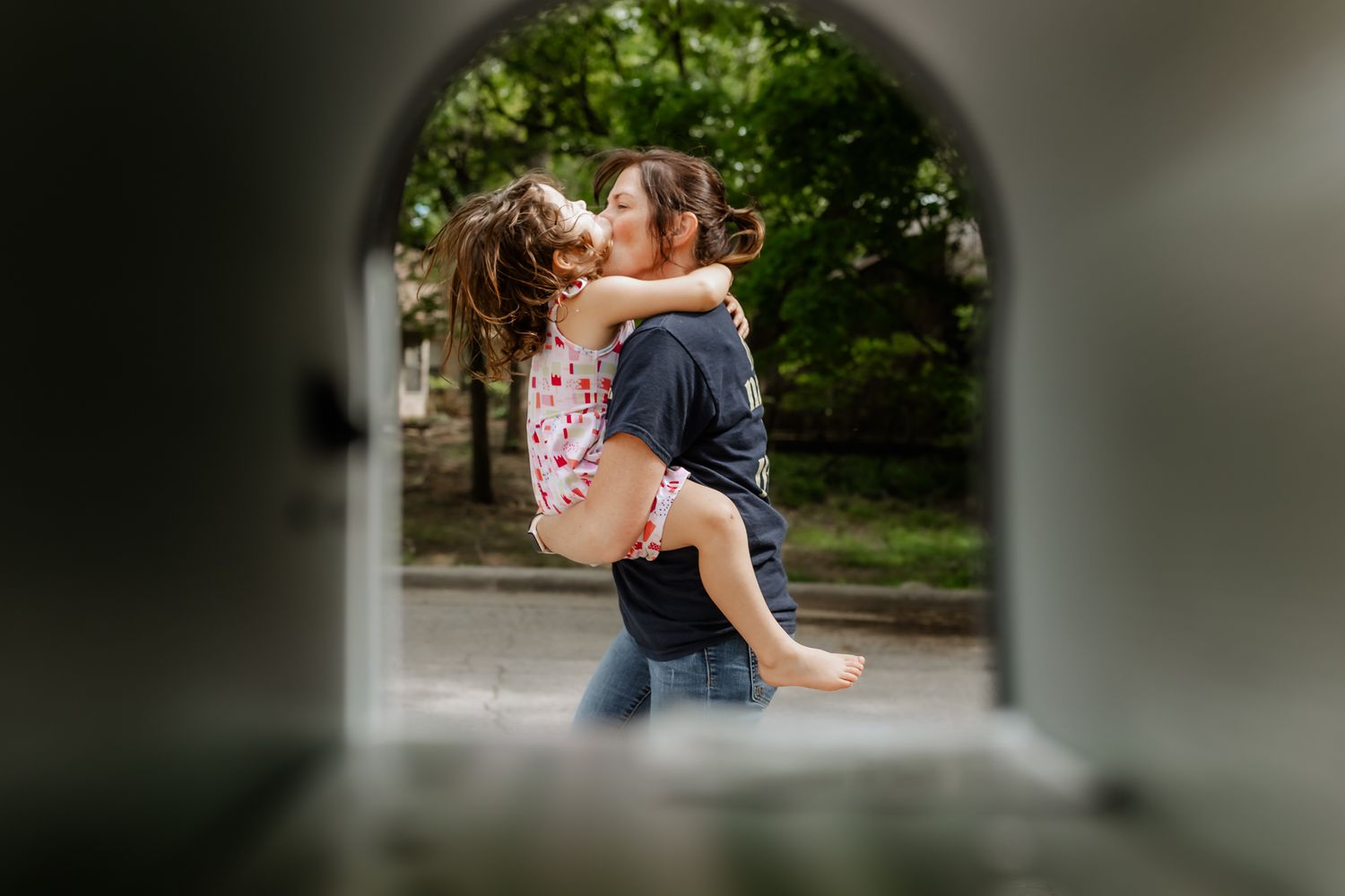 A woman joyfully lifts and hugs a young girl outdoors, framed through a mailbox a creative way to document families with Liz Perez Lawton.