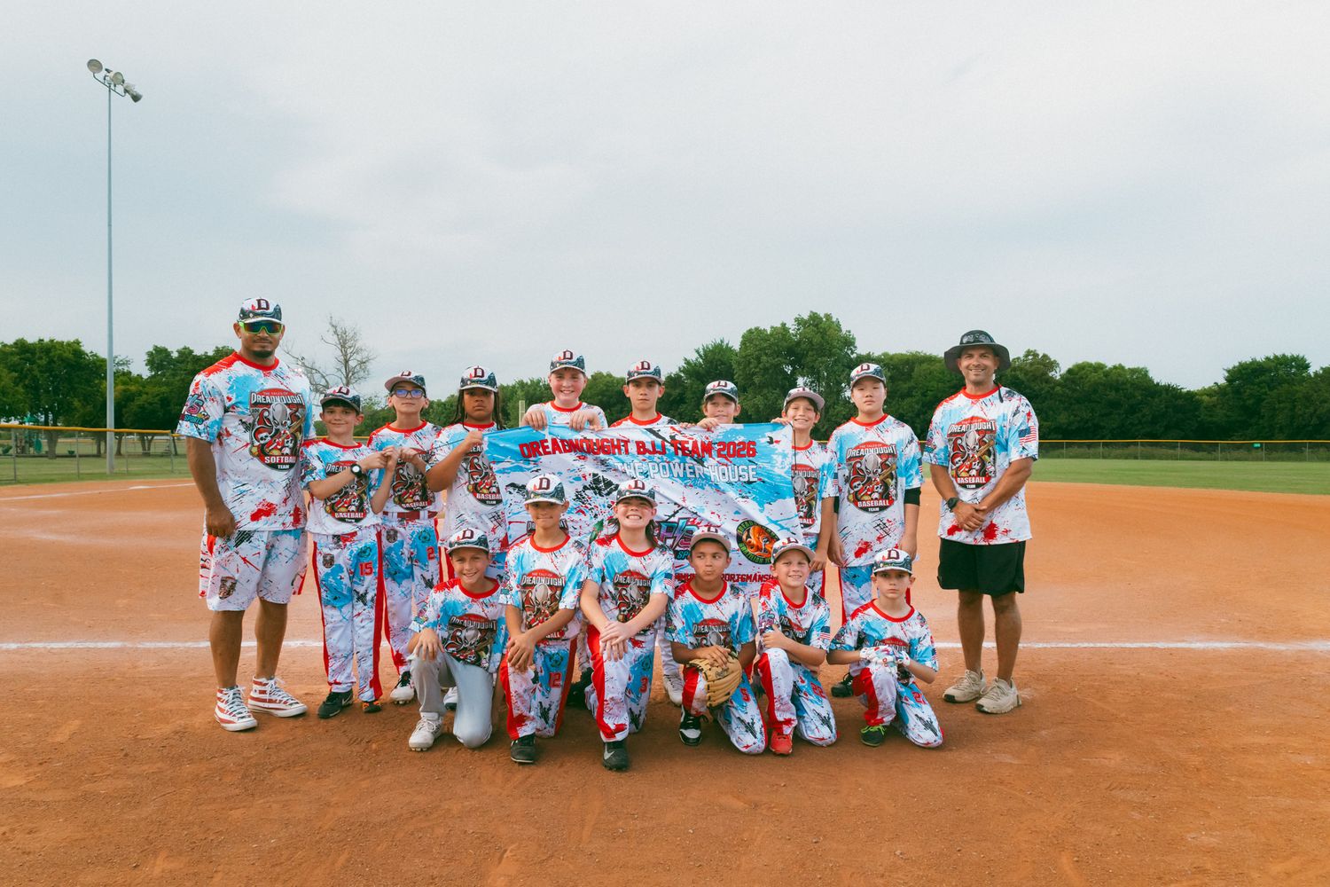 Youth baseball team posing together on dirt infield holding team banner after game.
