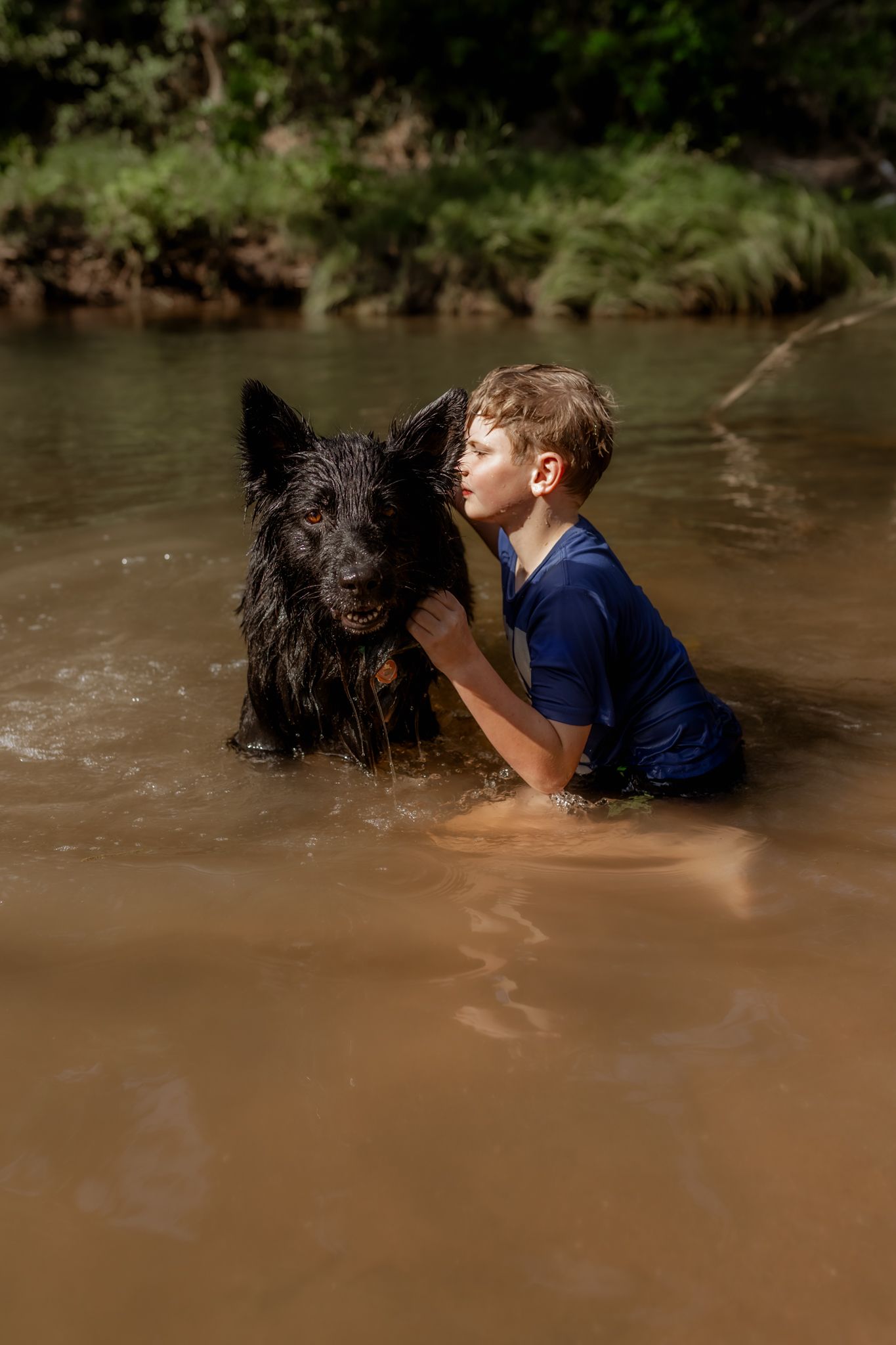 A wet black dog and child share a moment in murky river water.