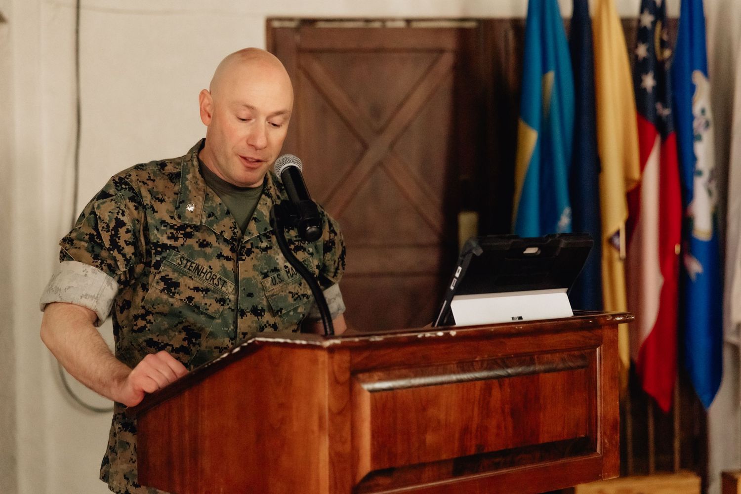 Military officer in camouflage uniform speaking at wooden podium with international flags displayed in background.