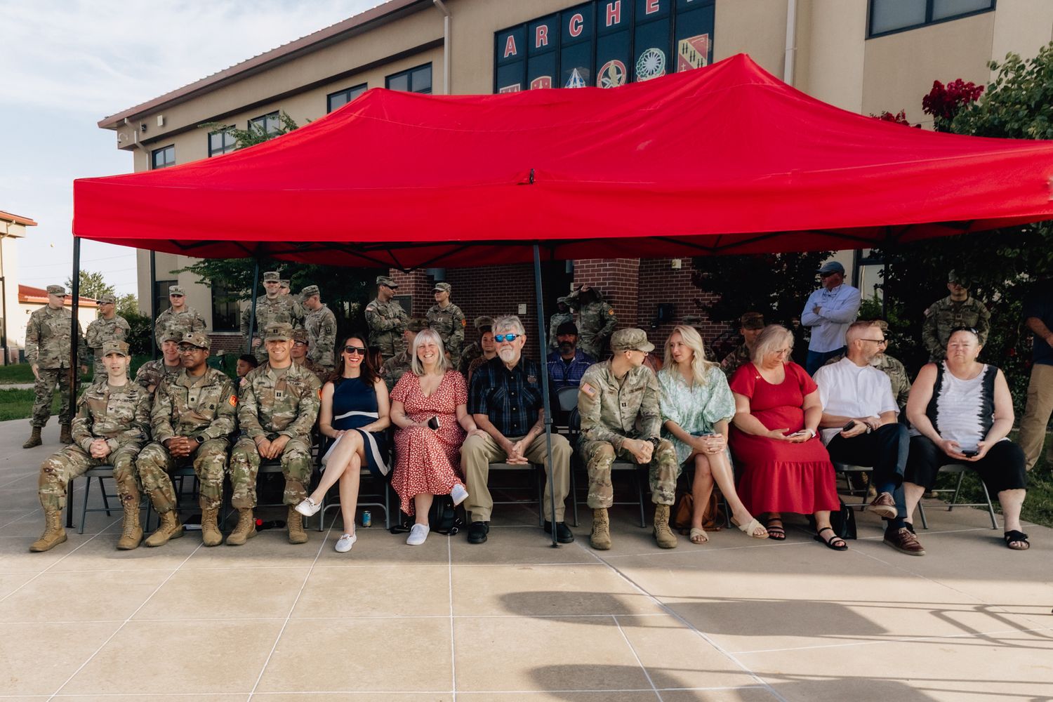 A group of people sit under a red canopy tent outside a building on a sunny day.