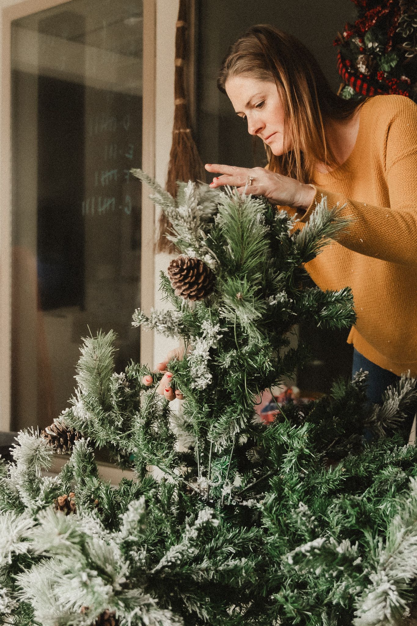 Woman arranging holiday greenery and decorations through a window.