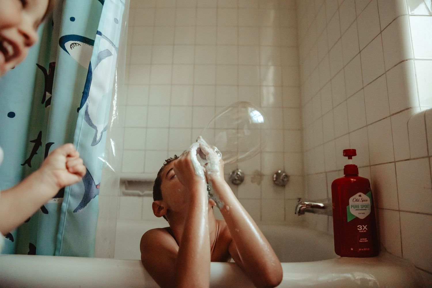 Child playing with bubbles in a bathtub with a colorful shower curtain and Old Spice shampoo nearby.