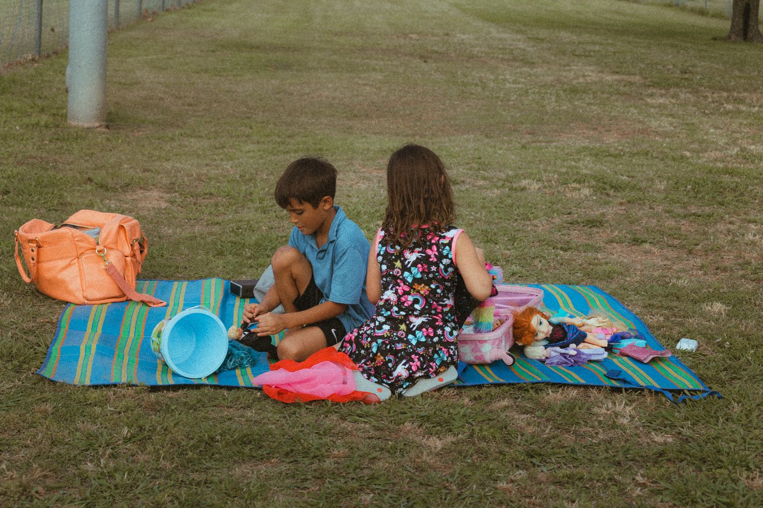 Two children sit on a striped blanket in a park, playing together with toys and a bag nearby.