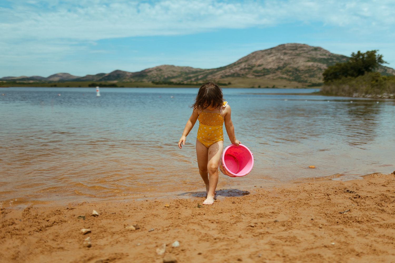 Child holding pink bucket walks along sandy beach shore near mountains and lake on sunny day.