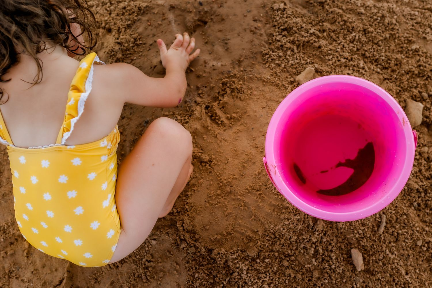 Child in yellow polka dot swimsuit playing in sand next to bright pink bucket.