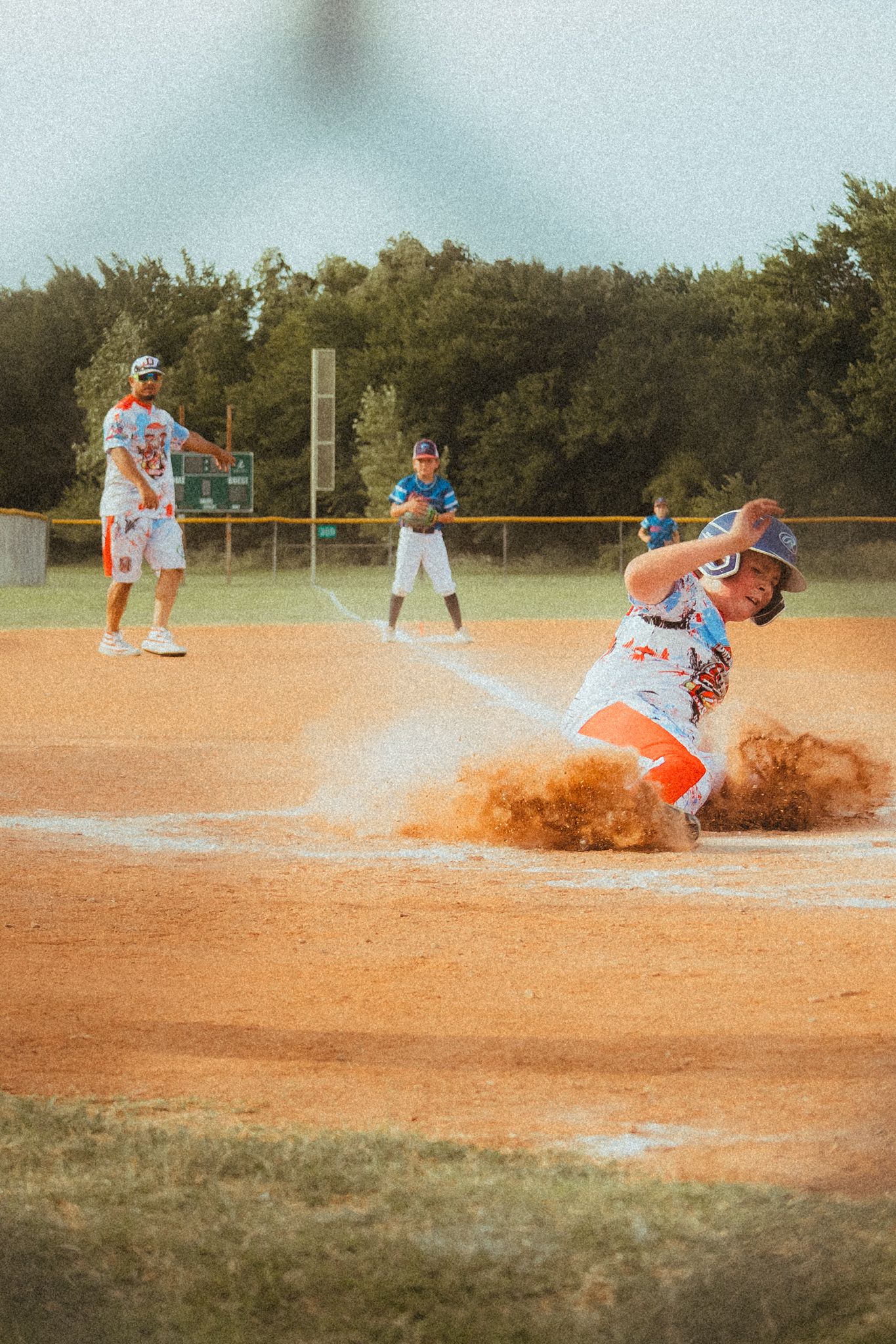 Baseball player sliding into home plate in a cloud of dirt during a youth league game on a sunny day.