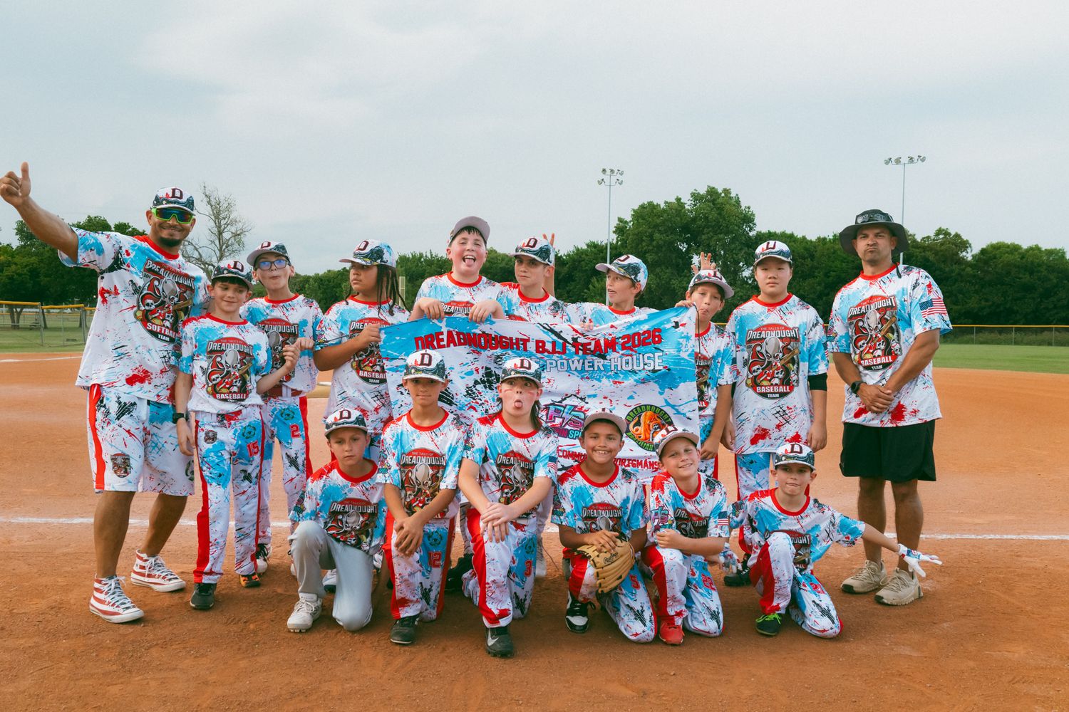 Youth baseball team poses together on field wearing matching red, white, and blue uniforms holding a championship banner.