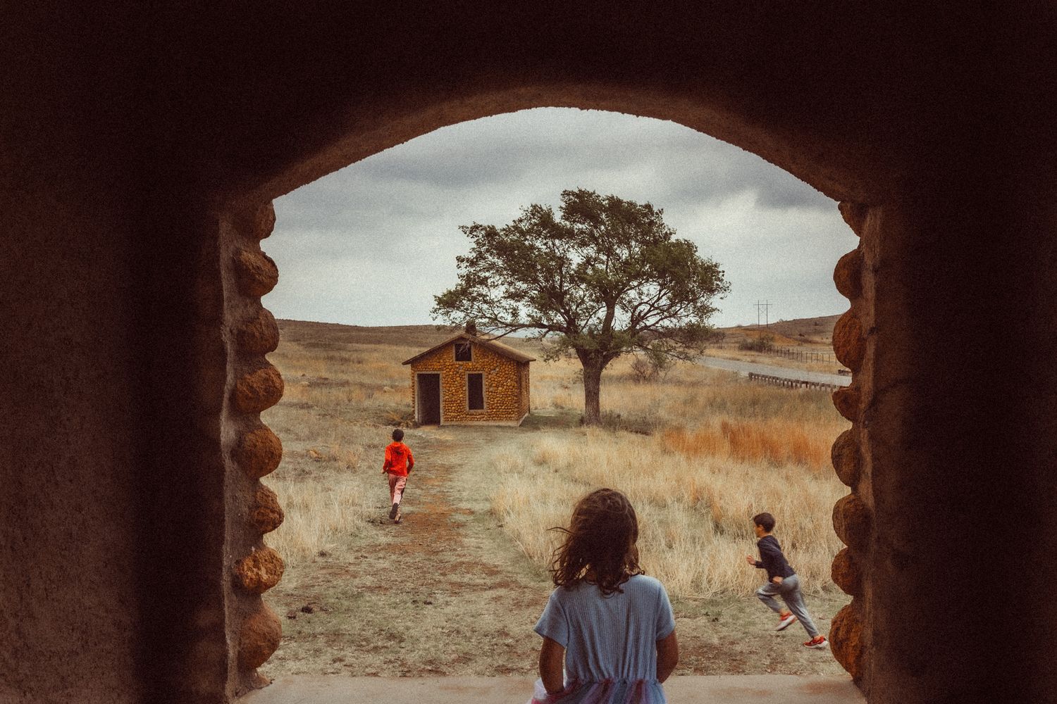 Children playing in an arid landscape viewed through a rustic stone archway with a wooden cabin and lone tree beyond.