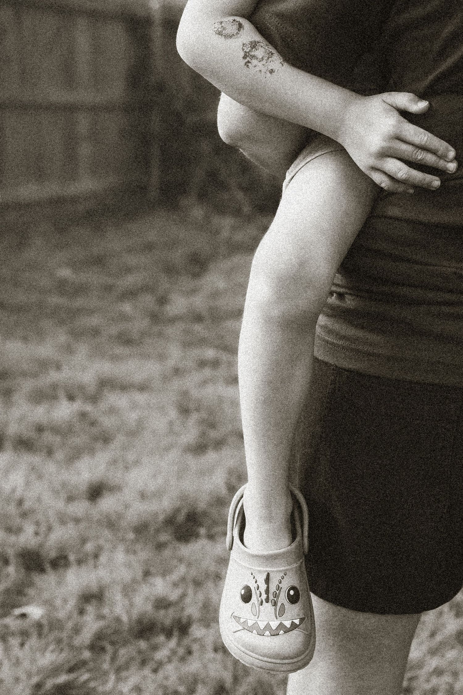 Black and white photo of adult holding child wearing monster Crocs shoes, with a small tattoo visible on the adult's arm.