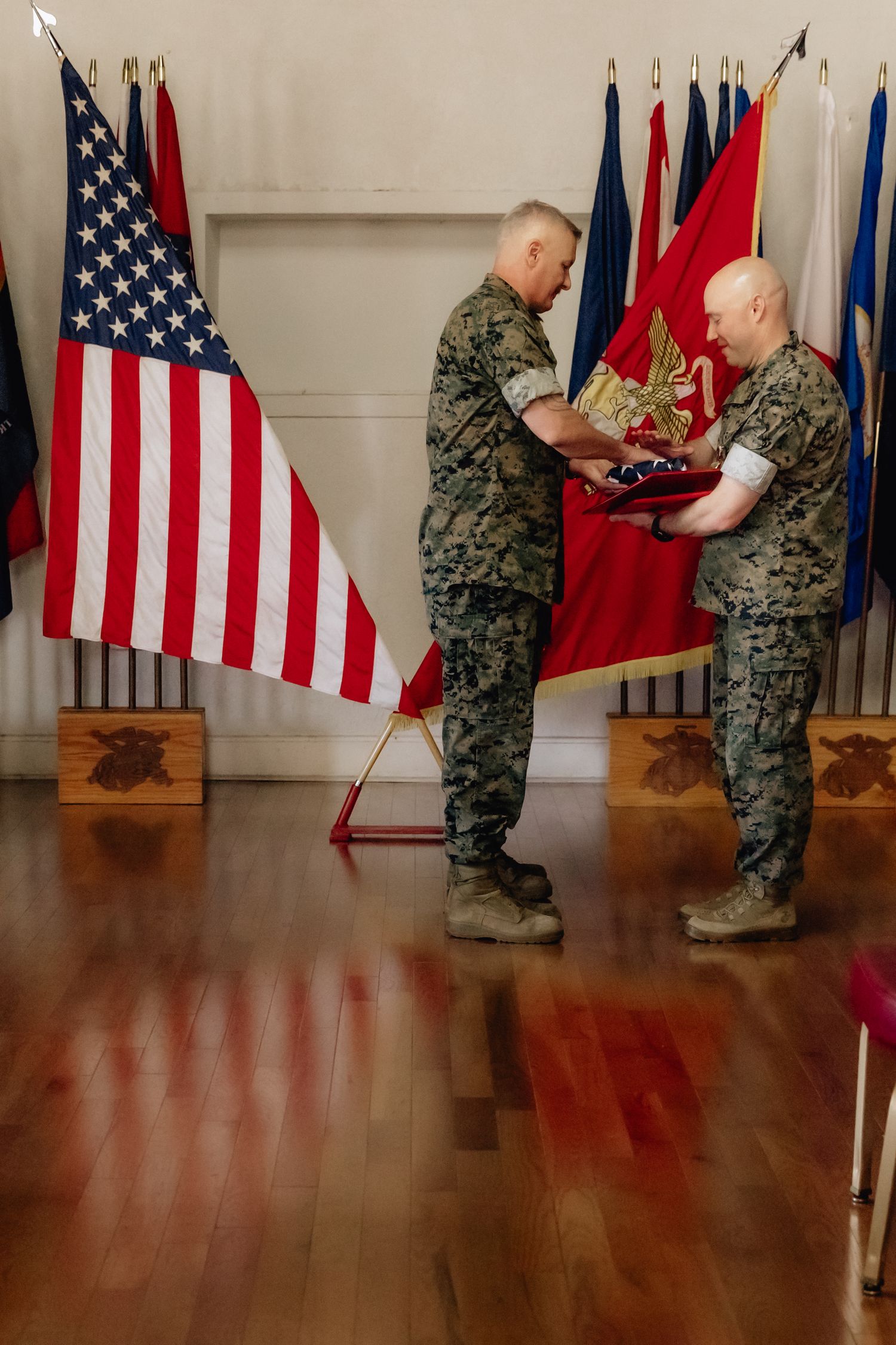 Two US Marines in camouflage uniforms exchange a folded flag during an official military retirement ceremony.