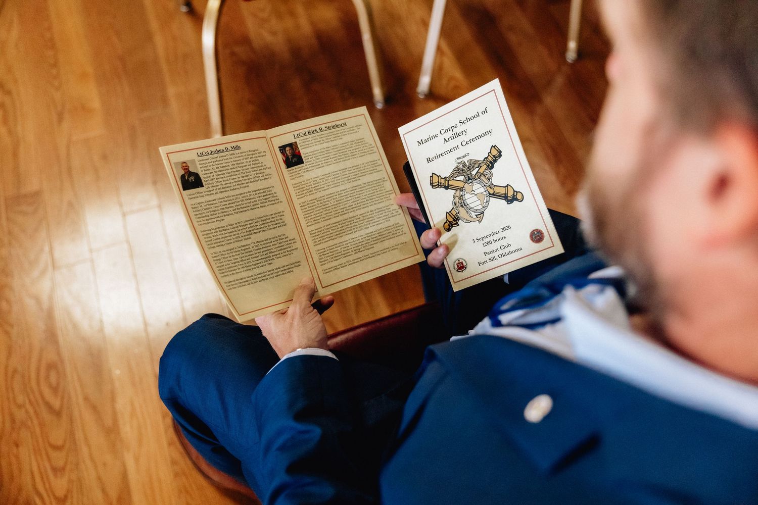 Man in blue suit reading a folded church program booklet while seated on wooden floor.