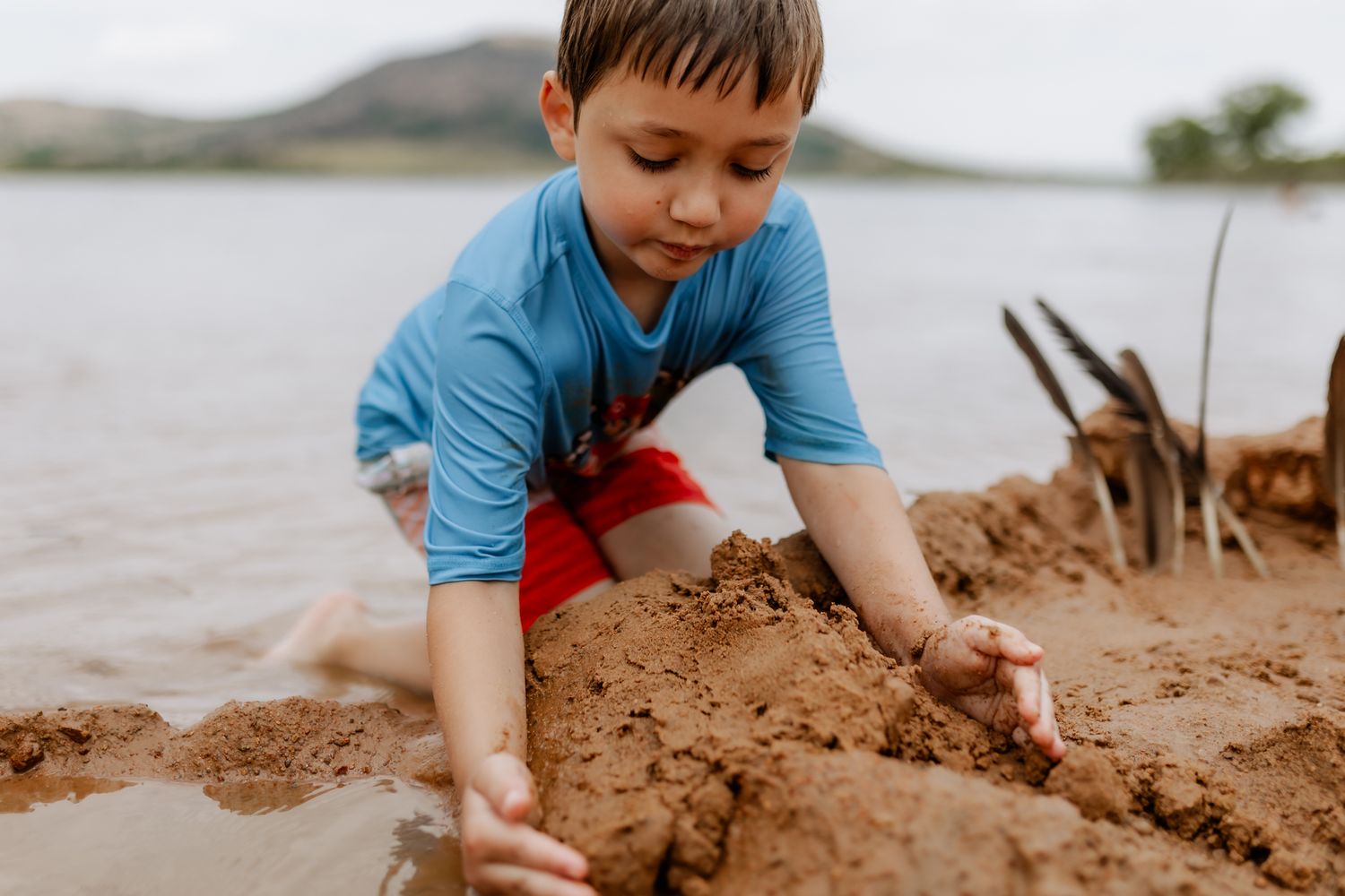 Child in blue shirt carefully molding sand formations by the water.
