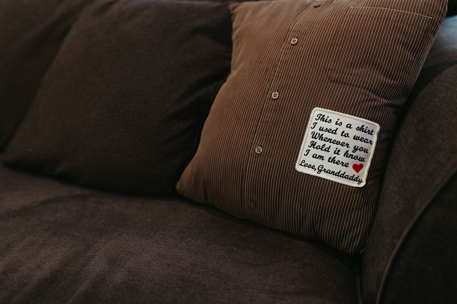Brown corduroy pillow with handwritten sentiment patch resting on a dark brown sofa.