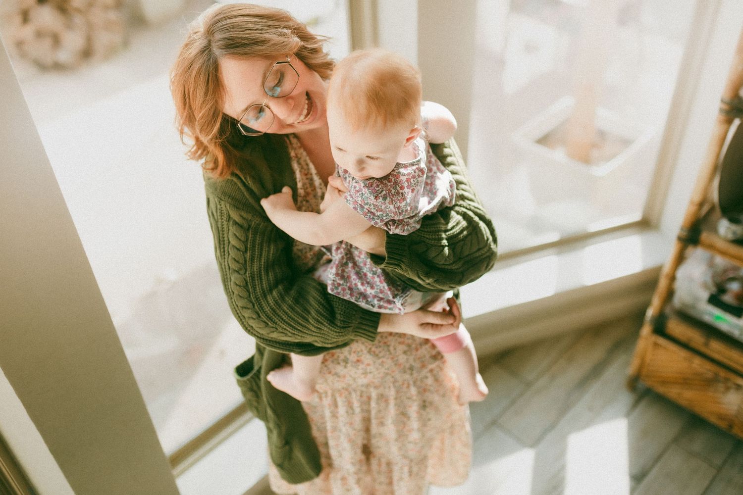 A parent in a green sweater holds a baby wearing a floral dress by a bright window.