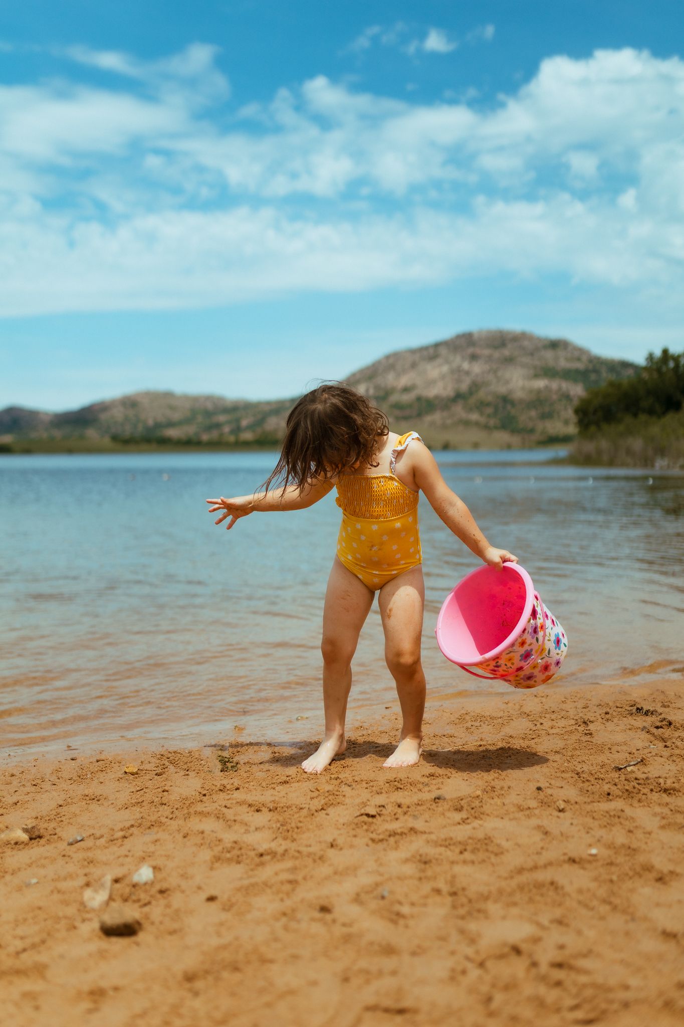 Child in yellow swimsuit playing with pink bucket on sandy beach near mountain lake.
