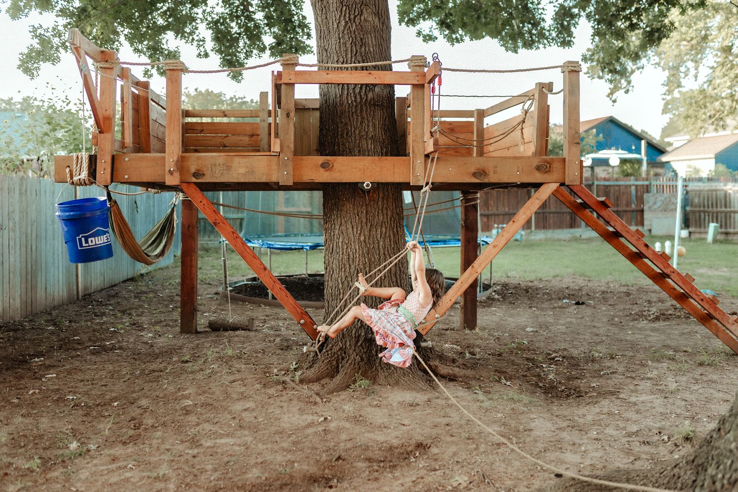 Wooden treehouse platform built around a large tree trunk with rope swings hanging underneath in a backyard setting.