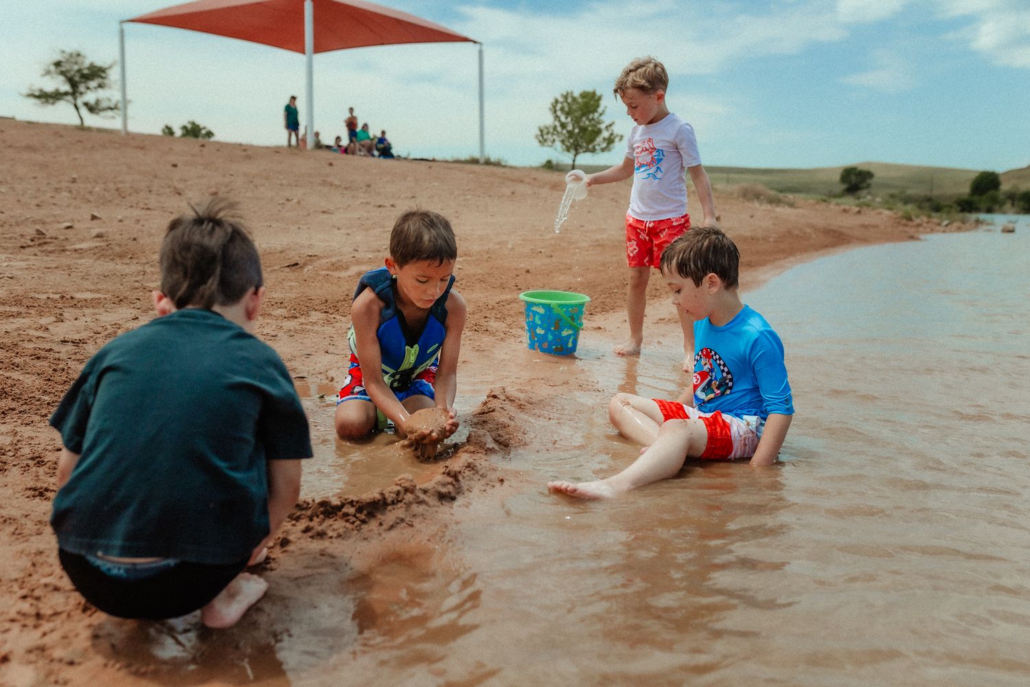 Group playing and digging in sand at water's edge under red sunshade.