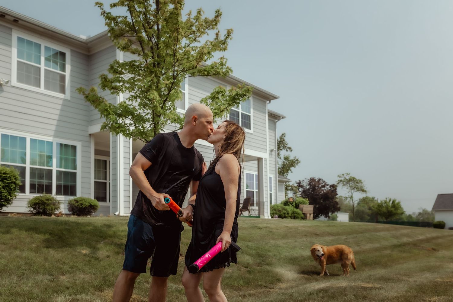 Couple playing with water guns in their backyard with a dog visible in the background.