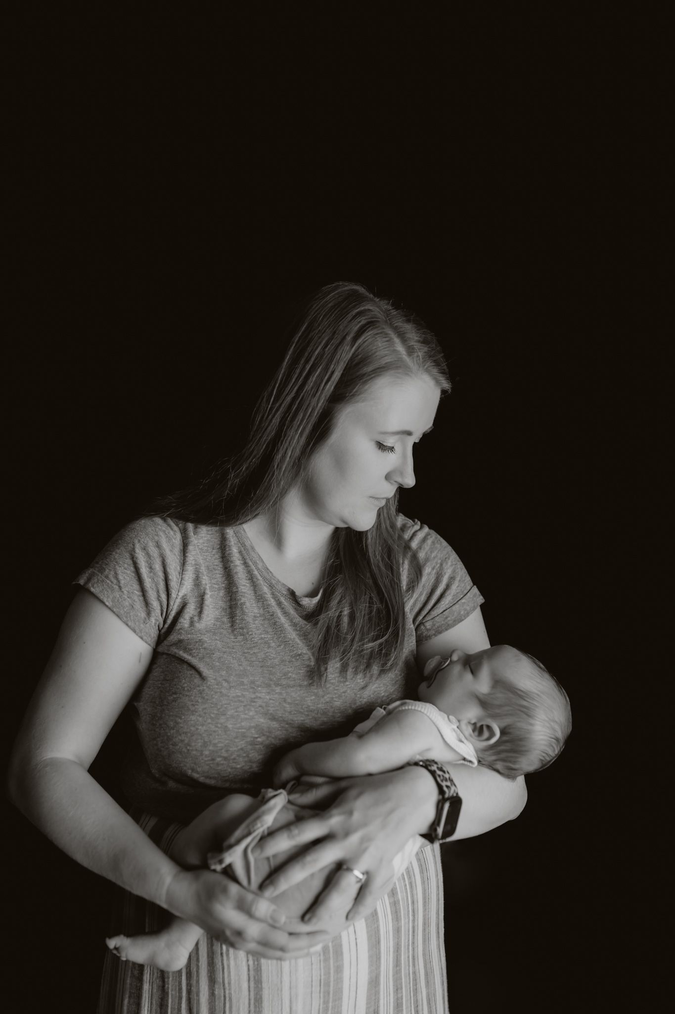 A tender black and white portrait of a mother in a gray shirt cradling a sleeping newborn baby against dark background.