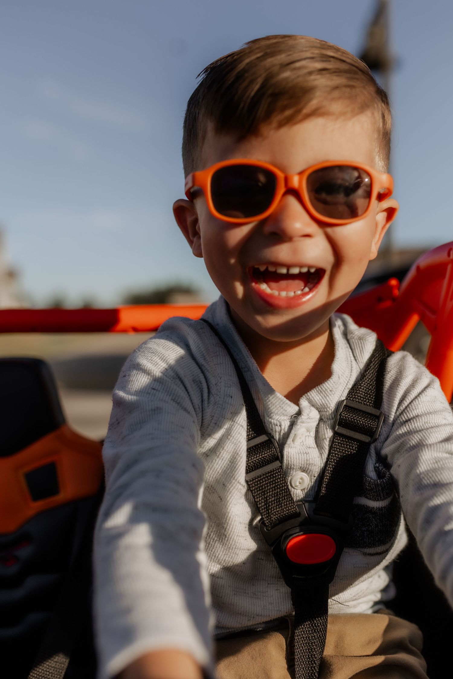 Young child wearing orange sunglasses smiles brightly while sitting in vehicle.