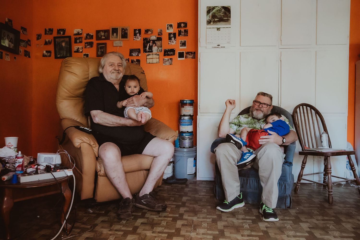 Two people sit in chairs in a room with orange walls covered in family photographs.
