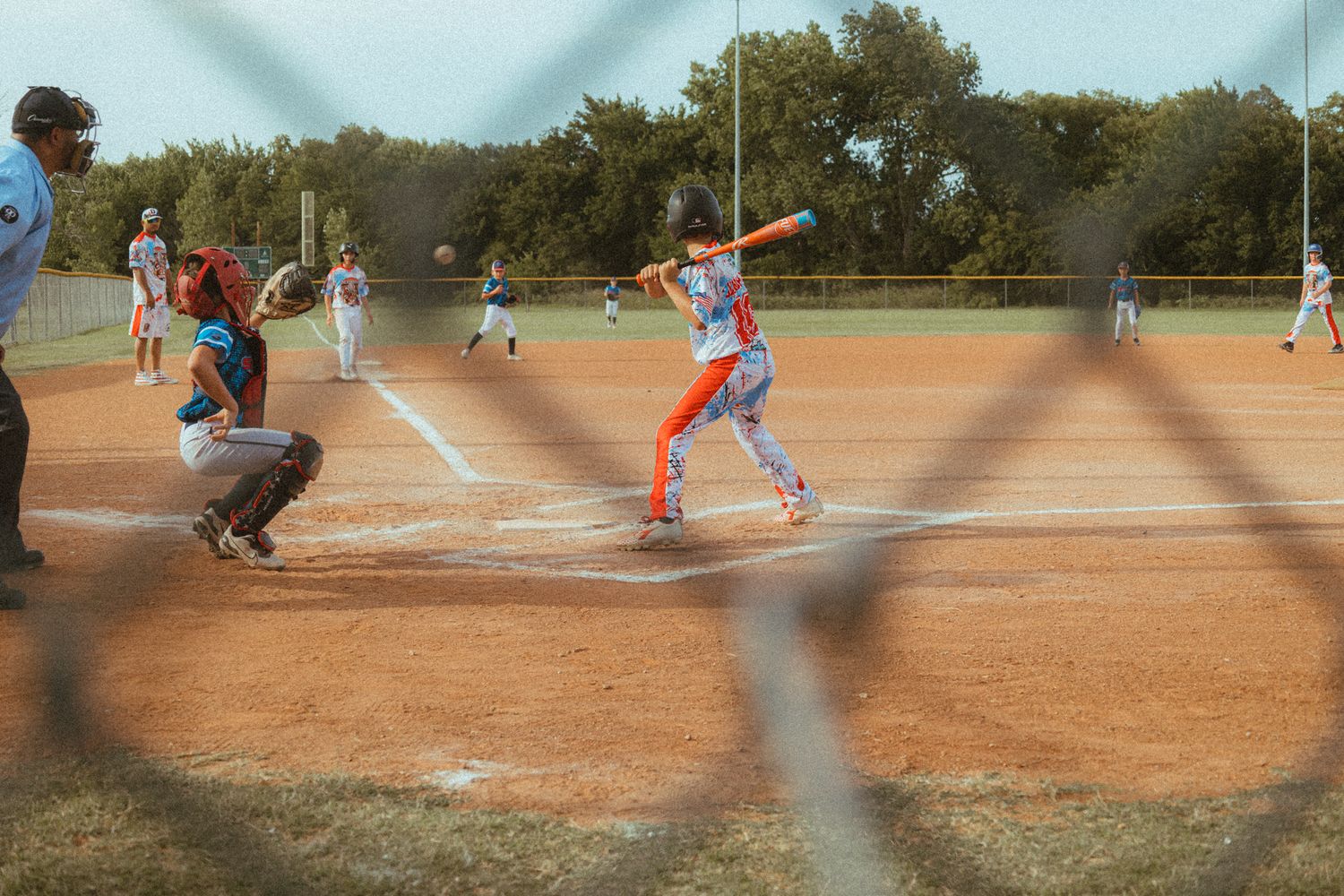 Youth baseball batter at home plate ready to swing, with catcher and umpire behind, viewed through chain-link fence.