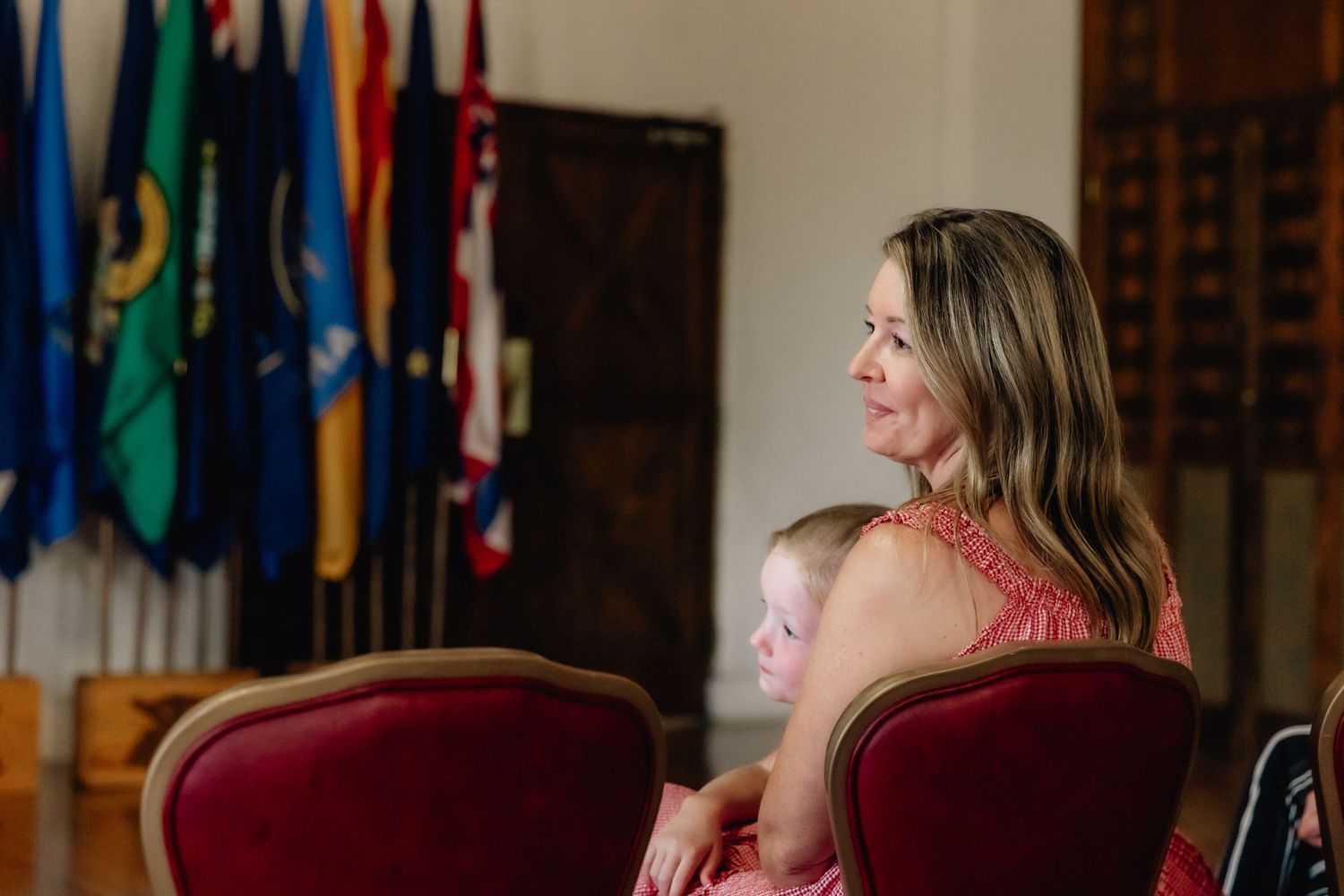 Woman and young child sit in red velvet chairs watching a military ceremony with colorful flags in background.