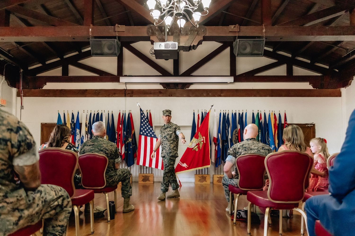 Military ceremony in a decorated hall with soldiers and civilians seated before flags and a speaker at the front.