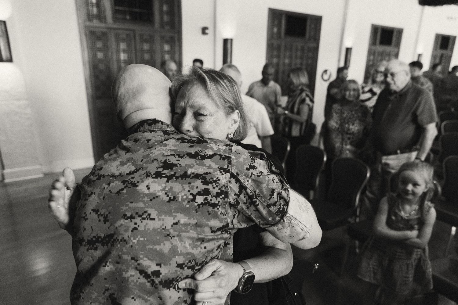 Black and white photo of a soldier in camouflage uniform sharing an emotional hug with a woman at a military homecoming event.