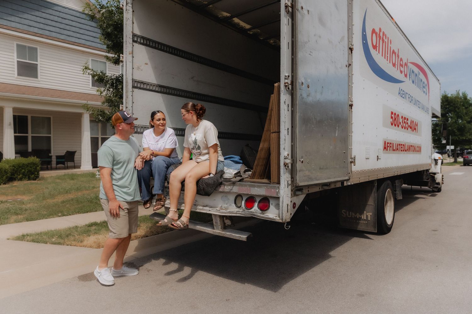 Three people relaxing on the back of an Affordable Moving truck outside a suburban home on a sunny day.
