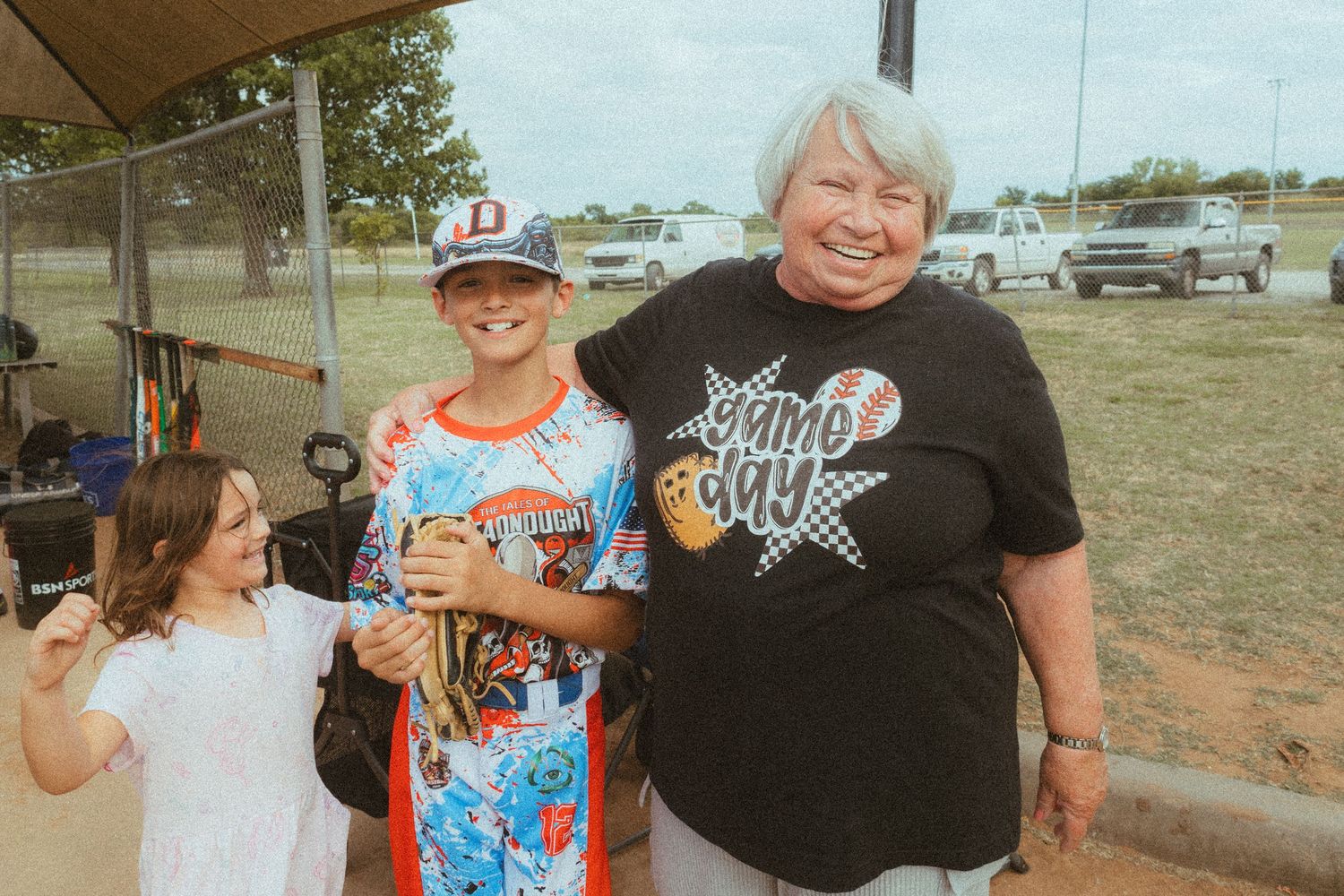 Three generations of family smiling together outdoors at a baseball field, wearing sports jerseys and game day shirts.