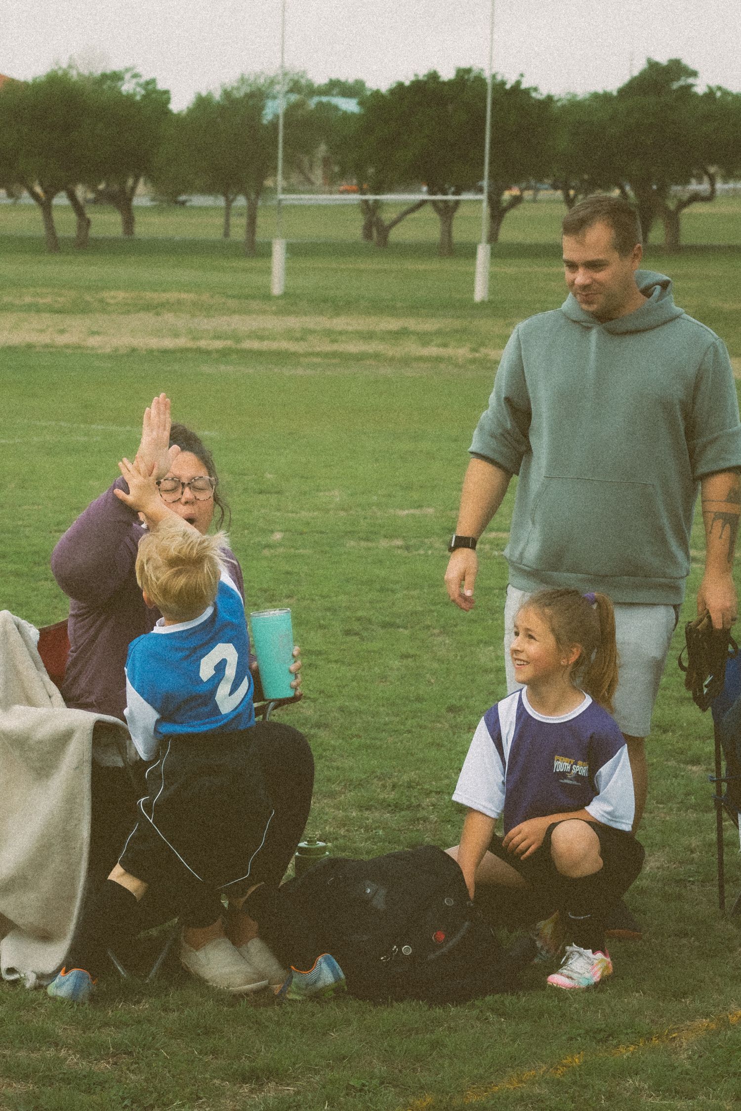 Adults and children in sports jerseys gather on a green field, with a woman giving a high-five to a young boy.